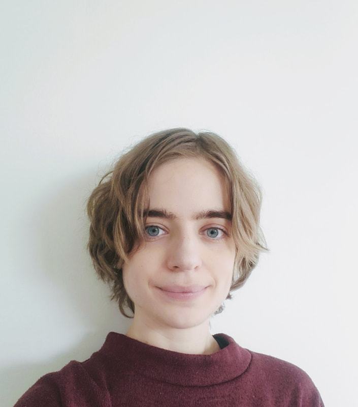 Veronique, a while non-binary person with wavy, sandy blonde hair falling just below their ears, stands with a white wall behind them. They are smiling a little and wearing a dark red crewneck sweater.