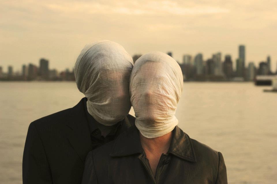 Two people wearing dark clothes stand close together, almost embracing. Behind them is a blurry cityscape. Their faces are concealed by swaths of white, semi-transparent fabric.