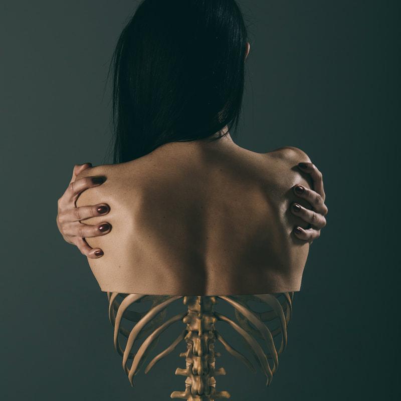 A dark-haired, brown-skinned woman, with her back turned, stands against a dark green background. A visual effect gives the impression that her flesh melts away, leaving the bones of her spine and ribcage.