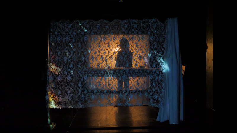 A performer speaks into a mic as they stand silhouetted behind a scrim and a lace curtain.