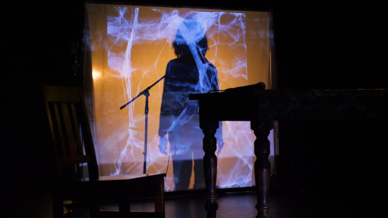 A performer stands silhouetted behind a scrim, speaking into a mic. A bluish, cracked texture is projected onto the scrim.