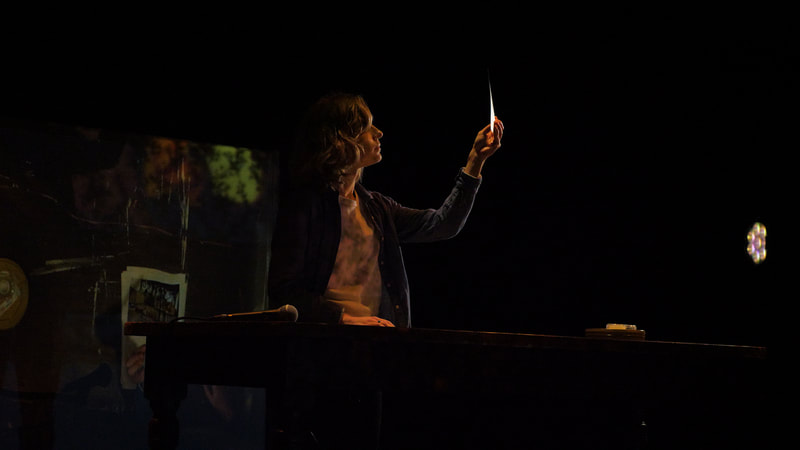 A performer stands at a table, lifting and inspecting a piece of paper. Behind them, a shadowy texture is projected onto a scrim.