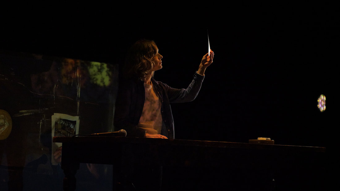 In a dark studio space, a performer stands behind a table, inspecting a document. Behind them, a shadowy texture is projected onto a scrim.