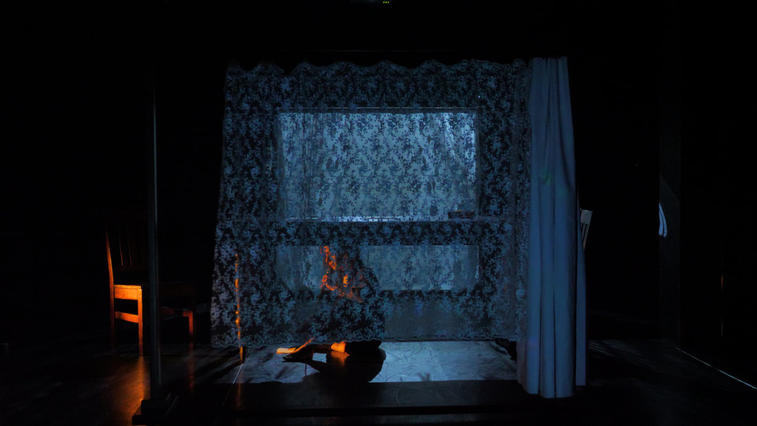 In a dark studio space, a performer sits under a table, speaking into a microphone. They are lit by a warm glow. A lace curtain hangs in front of the table.