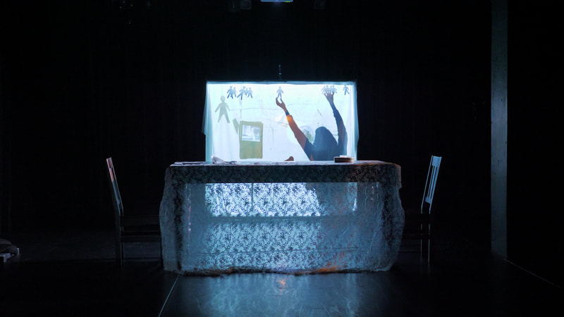 A performer stands silhouetted behind a scrim, arranging human-shaped shadow puppets. In front of the scrim, is a table with two chairs and a lace tablecloth.