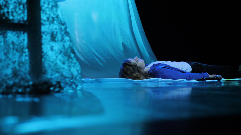 A performer lies on their back on the floor of a dark studio, bathed in blue light. Behind them, the blue light spills onto a white scrim.