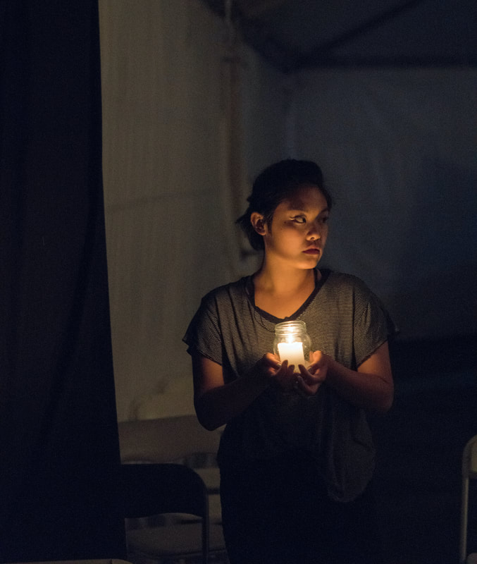 A woman of Chinese descent walks through a dark space, holding a lit candle in a glass jar. She glances to the right cautiously, her face glowing in the candlelight.