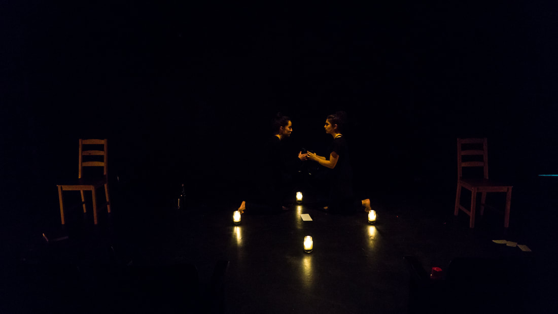 In a dark studio, two performers kneel facing each other and holding hands. A circle of lit candles in glass jars surrounds them.